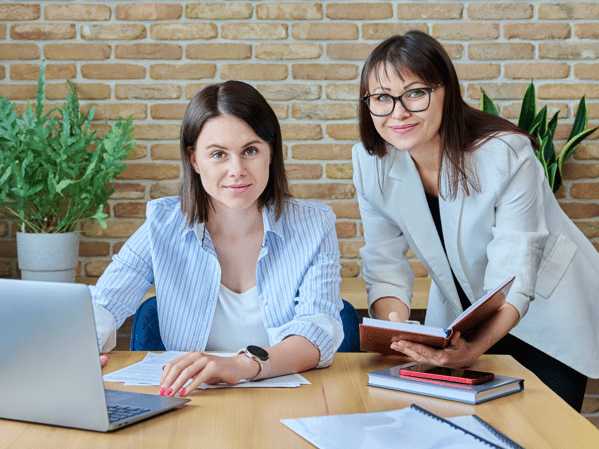 Two professional women working together at a desk, one seated with a laptop and papers, the other standing with a notebook, symbolizing collaboration and teamwork in a modern office setting.